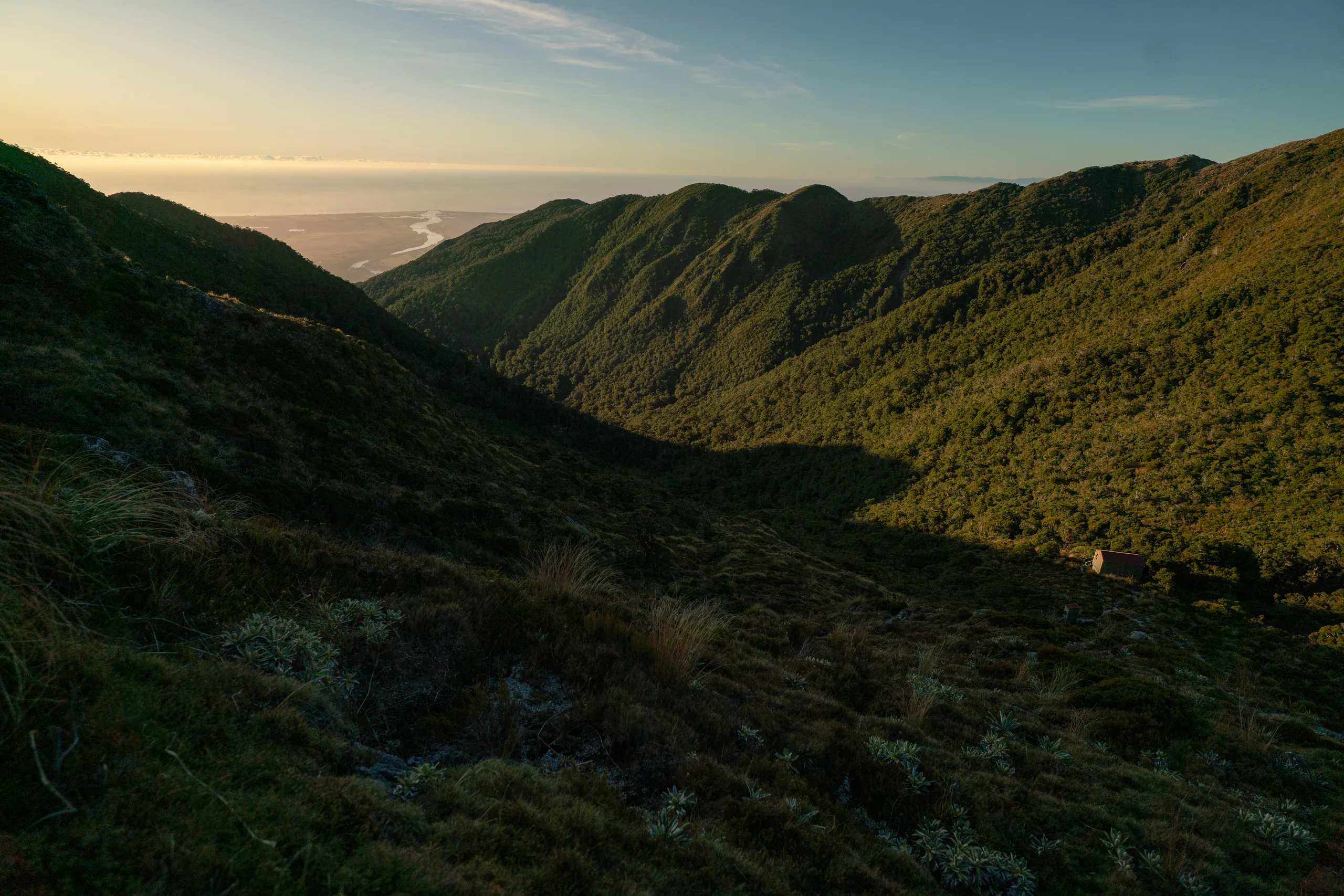 Buckland Peaks Hut as viewed from the saddle. The mouth of the Buller River is in the distance, framed between hills.