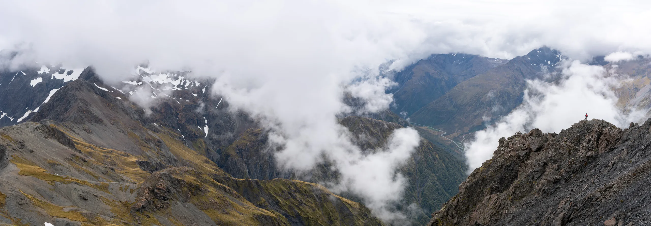 Dramatic vista from the summit. A hiker can be seen admiring the view. Far below, the highway winds up towards Arthur's Pass proper.